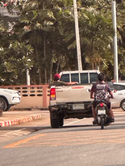 In Ban Thung Aok, Thailand, a white pickup truck with a beige cab is parked on a paved road. A person wearing a red cap is visible in the driver's seat of the pickup truck. Behind the pickup truck, a person is riding a dark-colored motorcycle. This person is wearing a dark t-shirt with a graphic design and is looking forward. The motorcycle has a license plate that reads "7465". In the background, lush green trees and a beige wall with a red trim are visible. The scene appears to be during the daytime, under natural light.