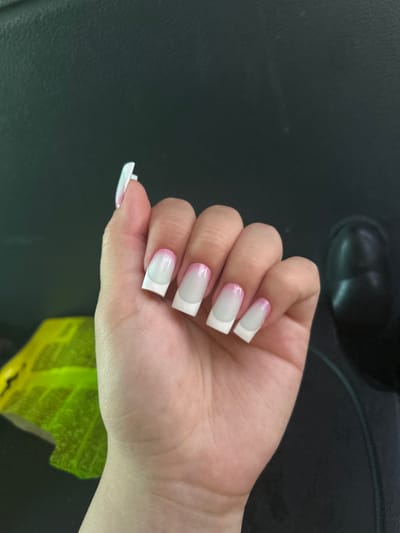A close-up, high-angle shot shows a left hand with acrylic nails that have a French manicure design. The nails are square-shaped with white tips and a gradient of pink and white on the base. The hand is positioned in front of a dark, textured surface, possibly a car interior or furniture. To the left of the hand, a bright yellow object with a leaf-like pattern is partially visible. The background is dark and out of focus. There is no identifiable location in the city of Maiaky, Ukraine, as the image is cropped and lacks contextual details.