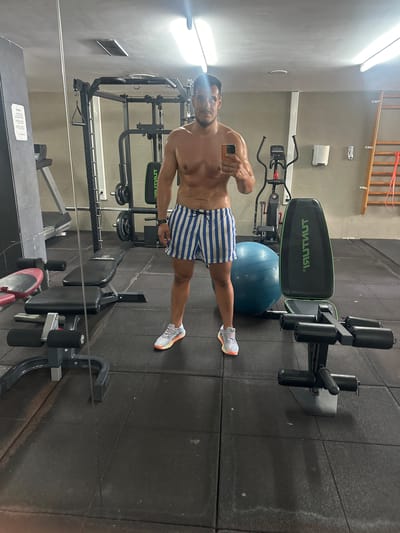 The image shows a man taking a mirror selfie in a gym. He is shirtless and wearing blue and white striped shorts and white athletic shoes. He is holding a smartphone in his left hand. Behind him, various gym equipment is visible, including weight machines, a weight bench with padded rollers, and a large blue exercise ball. The gym floor is made of black rubber tiles, and the walls are tiled with gray squares. A wooden rack is mounted on the wall in the upper background. The lighting suggests it is daytime, and the scene appears to be indoors. The overall mood is one of fitness and personal well-being. Visible text includes "ISITNUT" on the weight bench and some unreadable text on a document on a bench in the foreground. The location context is Roquetas de Mar, Spain.