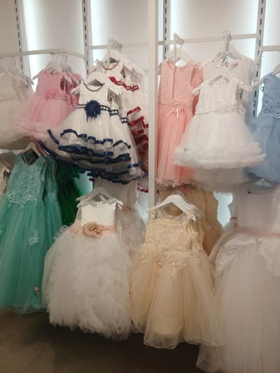 The image shows a display of formal dresses, likely for children, in a retail setting. The dresses are hung on racks and arranged in rows. They vary in color, including white, pink, blue, green, and a light peach or cream color. Several dresses are white with elaborate lace or tulle details, while others feature colorful ribbons or embellishments. The environment appears to be a brightly lit store with white vertical panels in the background. There are no people visible in the image, and no specific location within the city of Ávila, Spain, can be determined from the visual information provided. The focus is solely on the collection of dresses.