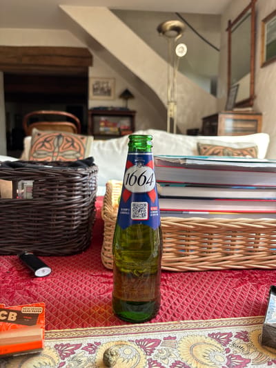 The image depicts an indoor residential scene in Bû, France. In the foreground, a green glass bottle of 1664 beer, approximately half-full, stands upright on a red quilted tablecloth with a decorative border. The bottle's label features a white "1664" logo on a blue background, accented with red, and includes a visible QR code.
To the immediate left of the beer bottle, a dark brown wicker basket is visible, partially obscured, with a black cylindrical lighter (white tip visible) resting horizontally in front of it. An orange and white packet labeled "OCB NEW DESIGN SAME PAPER VIRGIN Slim" is positioned in the lower-left corner. To the immediate right of the beer bottle, a lighter-colored wicker basket is present. A stack of multiple books with colorful spines is situated directly behind the beer bottle, between the two wicker baskets. A small, irregular, yellowish-brown object rests on the tablecloth's decorative border in the lower-center, and a metallic object, possibly an ashtray, is partially visible in the lower-right corner.
In the midground, a white sofa with several cushions, including one featuring an orange and brown abstract design, is positioned behind the table. The background reveals a living area. To the left, a prominent wooden ceiling beam is visible. Various pieces of furniture, including two chairs with woven backs and a display cabinet with contents, are present in a dimly lit alcove. To the right, a staircase ascends, and a tall, bronze-toned floor lamp with two light fixtures (one upward-facing, one forward-facing) stands. A wooden-framed mirror and a framed picture adorn the wall to the right. Natural light illuminates parts of the room from an unseen source. No individuals are present in the scene.
