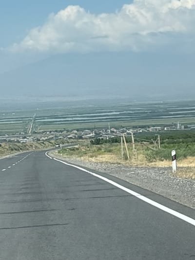 The image depicts a rural roadside scene in Paruyr Sevak, Armenia. A paved road with a white line marking curves to the right, bordered by gravel on the left. Beyond the road, a line of trees and structures suggests a small settlement, possibly a village. Fields, likely agricultural, are visible in the distance, characterized by parallel lines of cultivation or irrigation. A light blue sky with scattered white clouds indicates fair weather. The overall impression is of a peaceful, daytime scene in a rural landscape. The orientation of the image is vertical.