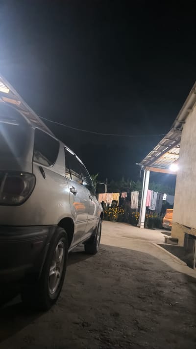 The image shows a silver SUV parked outdoors at night. The car is viewed from the rear passenger side, with its left rear wheel in focus. To the right of the car is a building with a covered outdoor area, illuminated by a bright light. Clothes are hanging on a clothesline in front of the building. In the background, there are trees and some yellow flowers. A portion of another vehicle, orange in color, is visible near the building. The ground appears to be a dirt or gravel surface. The sky is dark, indicating nighttime. The scene is likely a residential or rural area in Интернационал, Kyrgyzstan.