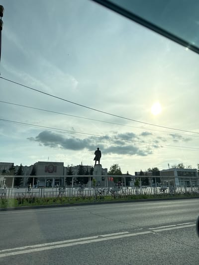 A street scene in Kstovo, Russia, includes a statue of Vladimir Lenin on a pedestal. It is partially obscured by clouds. The road is marked with white lines, and buildings with trees can be seen alongside it. The sky is bright, with the sun visible through the clouds. The photo was taken from inside a car.