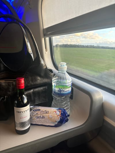 The image, taken from inside a train, shows a window with a view of green fields and cloudy skies. On the shelf next to the window are a bottle of Buxton water, a package of McVitie's Digestives White biscuits, and a bottle of Trivento Malbec wine. A black leather bag is also present, partially obscuring the items on the shelf. The image does not confirm that it's in Cheddington, United Kingdom.