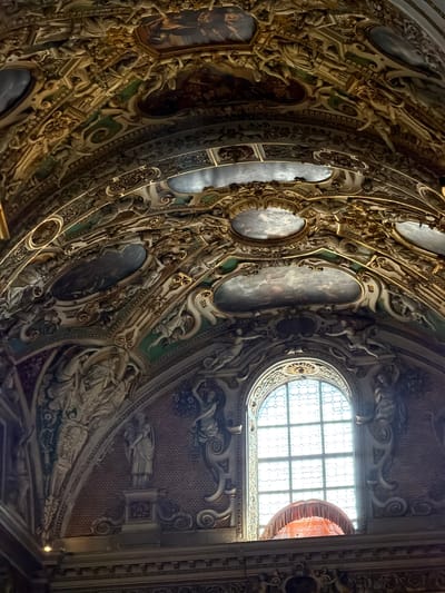 The image shows the interior of a richly decorated ceiling in a building located in Mozzo, Italy. The ceiling is adorned with intricate golden stucco work, featuring various ornamental patterns, scrolls, and sculptural elements. Several oval and rectangular panels depicting paintings of celestial or mythological scenes are embedded within the stucco. The architectural style appears Baroque or Rococo, characterized by its opulence and elaborate ornamentation.
Below the ceiling, a large arched window with a grid-like pattern is visible. The window is brightly lit from the outside, casting a soft glow. In front of the window, an ornate, draped fabric structure, possibly a canopy or curtain, is visible, adding a touch of color with its orange or pink hue. The walls surrounding the window are decorated with relief sculptures of figures, some appearing to be classical statues. The overall impression is one of grandeur and historical significance, indicative of a church, palace, or other important historical building.