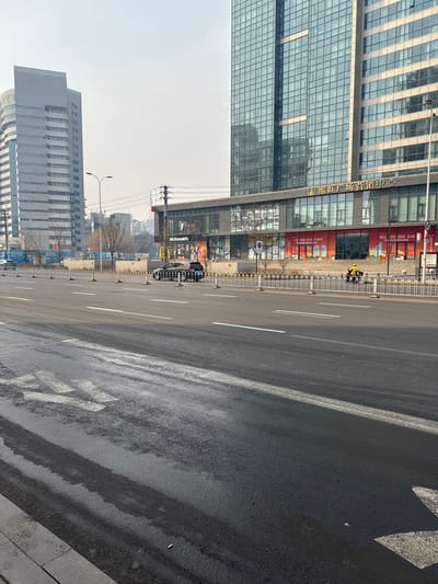 This image captures an urban street scene in Dadong District, Shenyang, China, under a daylight sky with slight atmospheric haze. The foreground features a multi-lane asphalt road with visible moisture or residue, exhibiting tire tracks and white lane markings, including a faded left-turn arrow painted on the surface in the lower-left quadrant.

A low, white metal barrier with brown-capped posts separates the road from a paved area in the midground. To the right of this barrier, a dark-colored SUV is present, appearing either parked or moving slowly. Further right, a person clad in a bright yellow jacket operates a yellow scooter on an adjacent lane, moving towards the right side of the frame.

The background is dominated by two large multi-story buildings. On the right, a tall, modern skyscraper with a reflective glass facade features a ground-level commercial complex. A "McDonald's" sign with its golden arches is prominently displayed, alongside other retail units showcasing large red promotional banners. Above the ground-level businesses, Chinese characters "嘉熙里广场营销中心" (Jiaxi Li Plaza Marketing Center) are visible. To the left, another tall, rectangular building with a gray, less reflective facade and numerous windows stands. Sparse, leafless trees are interspersed between the street and the buildings. Utility poles with overhead wires are also visible against the sky.