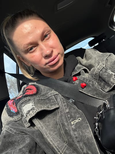 A person is positioned in the driver's seat of a car, wearing a gray denim jacket with red heart-shaped pins and a sequined design, and a black top. They have light skin, blonde hair, and facial piercings. The car is positioned in an unidentified location, not identifiable as Pesyanka, Russia.