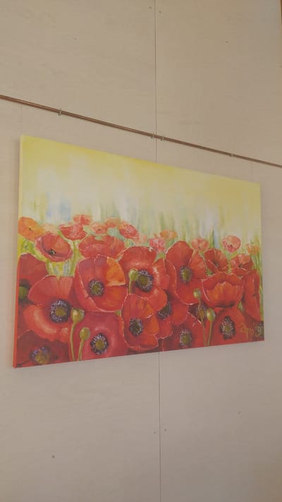 A painting of red poppies is displayed on a pale wooden wall in Tērvete, Latvia. The painting features a field of red poppies with yellow and green backgrounds. The poppies vary in size and stage of bloom, with some showing closed buds and others fully open petals. The artist's signature, "App 19," is visible in the bottom right corner of the painting. The painting is hung using a copper rod attached to the wall with metal hooks. The wooden wall is divided by vertical seams and has visible nail holes.
