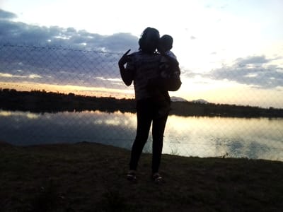 The image depicts an outdoor scene in Central Kasipul ward, Kenya, during either dawn or dusk. In the foreground, two individuals are silhouetted against a luminous sky. One adult individual stands centrally, facing slightly towards the right, holding a young child against their left hip. The adult's right hand is raised, with two fingers extended in a "V" gesture. Both individuals are rendered in near-total shadow, obscuring specific facial or clothing details beyond general shapes. A chain-link or wire mesh fence is positioned horizontally across the mid-ground, separating the individuals from a body of calm water that reflects the sky. The foreground consists of dark, uneven terrain, likely earth or sparse vegetation. Beyond the water, a dark, indistinct landmass with subtle hills or mountains is visible on the distant horizon. The sky exhibits a gradient of colors, transitioning from bright yellow and orange near the horizon line to lighter blues and grays higher up, with fragmented clouds present.