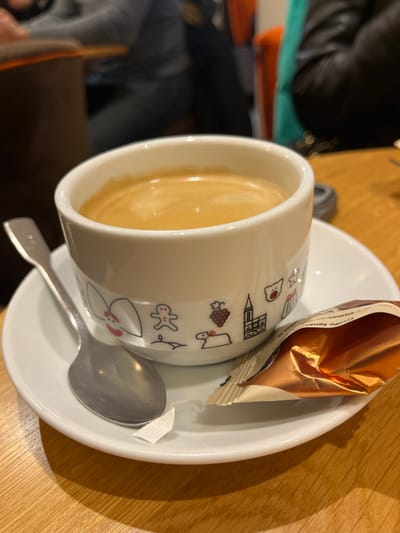 A white ceramic espresso cup containing coffee with a light brown crema is centered on a white saucer. The cup's lower circumference is decorated with a band of black and red outlined symbols, including a red-capped figure, a gingerbread man, a candle, red grapes, a church-like building silhouette, a lamb, a bear head, a star, and a cake. A silver-colored spoon rests on the saucer to the left of the cup. To the right, a partially unwrapped metallic bronze-gold foil package, visibly marked "truffle fantaisie," lies on the saucer, accompanied by a small, torn white paper fragment. These items are positioned on a light brown wooden table with a visible grain. The background is blurred, showing indistinct figures of at least two individuals, one in a dark jacket and another in a grey garment, within an indoor establishment indicated by abstract furniture elements, including a muted orange chair back. The scene is located in Sélestat, France, with the cup's iconography possibly representing local heritage or seasonal themes.