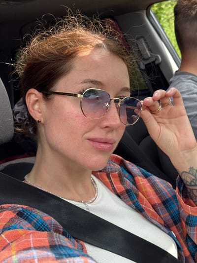 This media file shows a close-up, high-angle shot of a young woman and part of another person. The woman is lying down, likely in the passenger seat of a car, as indicated by a seatbelt. She is wearing large, aviator-style sunglasses with gold frames and tinted lenses. Her hair is a reddish-brown color and appears somewhat messy and curly, with some strands catching the light. She is also wearing a white top and an orange and blue plaid shirt over it. A silver necklace is visible around her neck. She has a small, dark red heart-shaped stud earring in her left ear.
The hand of another person, likely male given the visible tattoo on the forearm and the glimpse of a gray t-shirt, is holding the woman's sunglasses. This hand is adorned with two gold rings. The background shows the interior of a car, with a black seat and a gray headrest or armrest. Outside the car window, a patch of green grass is visible, suggesting an outdoor setting during the daytime. The lighting suggests it is sunny. The woman's facial expression is relaxed, with her eyes closed or looking down, and her lips are slightly parted. The overall mood is casual and possibly a moment of rest or enjoyment during a car journey. There is no visible text.