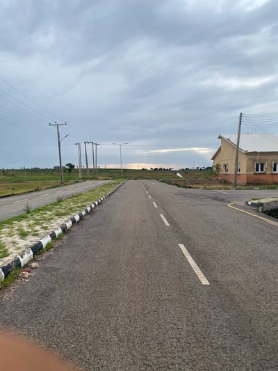 The image depicts an empty asphalt road with white dashed markings, curving towards a distant horizon. The road is bordered on the left by a strip of grass and a black and white striped curb. In the distance, on the right side of the road, a two-story building with a cream-colored facade and a white roof is visible, alongside a utility pole with electrical wires. The scene is set under an overcast sky, suggesting a cloudy or possibly a pre-rainy day. The presence of utility poles and the architecture of the building hint at a developed or developing residential area in Nigeria, likely in Bulamare given the location context. There are no people or specific activities visible in the image, giving it a tranquil, perhaps slightly isolated feel. The lighting suggests it is either early morning or late afternoon.