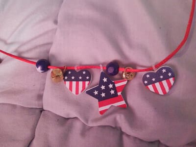 The image shows a close-up of a red cord necklace adorned with charms. The charms include several blue beads with white stars, and flat, wooden cutouts shaped like hearts and a star, all decorated with the American flag pattern of red and white stripes and a blue field with white stars. There are also small, gold-colored bell charms interspersed between the other charms. The necklace is laid out on a wrinkled, light-colored fabric, possibly a bedsheet or clothing. The lighting is soft, and the background is out of focus. The specific location in the city of Inhulo-Kamianka, Ukraine, cannot be determined from the visual information provided in the image.