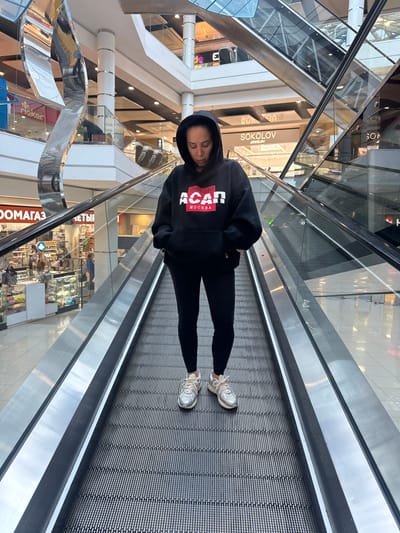 This media file shows a person standing on an escalator in a shopping mall. The person is wearing a black hoodie with red and white lettering that reads "ACAN МОСКВА" and black leggings. They are also wearing white athletic shoes. The escalator is moving upwards, and the background shows a multi-level shopping mall with various stores visible, including "SOKOLOV JEWELRY" and "rieker." The lighting suggests it is daytime. The person's head is tilted down, and their eyes are closed, giving an impression of relaxation or contemplation. The overall setting is a modern, well-lit indoor shopping environment. Text visible in the image includes store names and some Russian words.