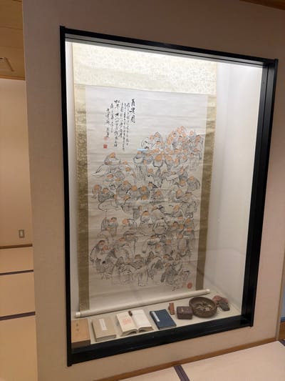The scene depicts an exhibit within an interior space in Yamanouchi, Japan. A large vertical scroll painting is prominently displayed inside a black-framed glass case that is internally illuminated from above. The scroll features numerous stylized figures, rendered in ink with accents of light orange or peach on their heads, gathered in a dense crowd. Calligraphy and red seals are visible in the upper left portion of the artwork. The scroll itself is bordered by patterned fabric in shades of light gold. Below the scroll, within the same display case, a collection of artifacts is arranged on a shelf. From left to right, these include a light-colored wooden box, two bound books (one closed, one open), a dark blue bound book, a small rectangular wooden block with red marks, a dark-toned small wooden box, a metallic bowl containing various small objects, and an irregularly shaped dark object. The display case is set against a light-colored wall. The floor in the foreground and extending to the left is covered with traditional Japanese tatami mats. An electrical outlet is visible on the wall to the left of the display case. No individuals are present in the immediate vicinity of the exhibit.