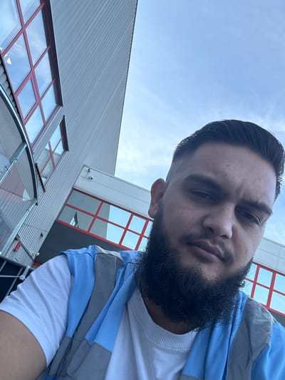 A low-angle selfie is taken outdoors, with the subject's face dominating the foreground. The man has dark hair, a full beard, and is wearing a white t-shirt layered with a blue and grey vest. In the background, a modern building with ribbed metal siding and large windows with red framing is visible. The sky above is clear and blue. The location is the city of Boom, Belgium.