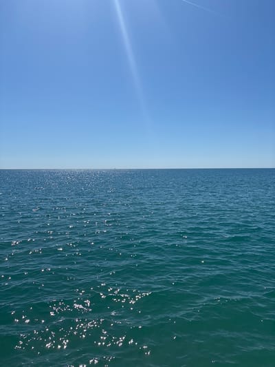 The image shows a stark division between a clear blue sky on the left and the surface of the Mediterranean Sea on the right. The sky is mostly featureless, except for a faint contrail in the upper left corner. The sea surface is rippled with small waves reflecting sunlight, creating a sparkling effect. The water appears dark blue-green. The location is likely off the coast of Fréjus, France, as the user specified. No people or other objects are visible. The image captures a simple contrast between sky and sea on a sunny day.