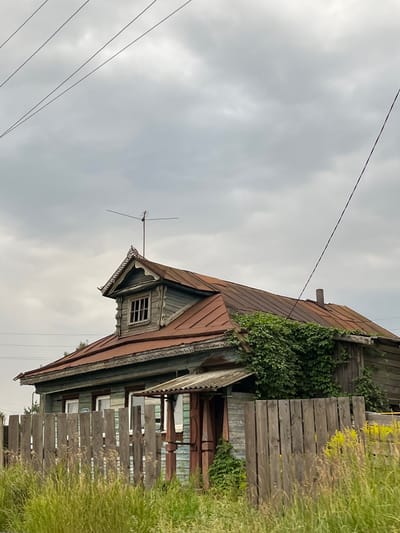 A weathered, two-story wooden house stands under a cloudy sky in Syrokhvatovo (Kantaurovskii S/S), Russia. The house has a rusted, reddish-brown metal roof with a dormer window on the left side, featuring decorative wooden trim. An old television antenna is mounted on the roof. Green vines climb up the side of the house and over a section of the roof. A wooden fence made of vertical planks encloses the property in the foreground, with tall green grass and some yellow flowers growing in front of it. Power lines are visible against the sky. The scene appears quiet and rural.