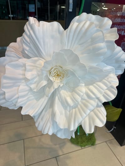 The image shows a close-up of a large, artificial white flower with many layered petals. The flower has a textured center with yellow stamen-like structures. It appears to be a decorative item, possibly in a shop or event setting. The background is blurred, showing a tiled floor and reflections in a window, suggesting an indoor environment. There is no information in the image to indicate it is located in Slitu, Norway.