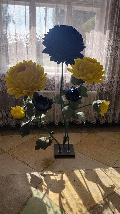 A large, artificial flower arrangement is displayed indoors. The arrangement features several large, multi-petaled flowers in shades of yellow and dark blue, with green leaves and stems. The flowers are mounted on a dark base. The arrangement is positioned in front of a window with sheer curtains, which are partially drawn. Sunlight streams through the window, casting shadows of the flowers and their leaves onto the tiled floor. The floor is made of large, square tiles in various shades of beige, yellow, and green. The image appears to have been taken on a sunny day. There are no people visible in the image.