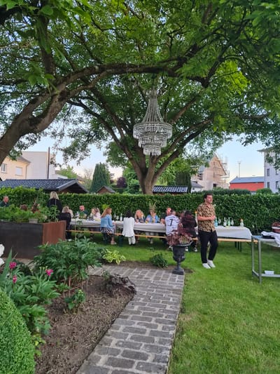 In Bettembourg, Luxembourg, an outdoor dinner party is taking place in a lush green garden. Several people are seated at long tables covered with white tablecloths, enjoying a meal under the canopy of a large tree. A crystal chandelier hangs from a branch above the tables, adding a touch of elegance to the setting. In the foreground, a meticulously maintained garden bed features vibrant green foliage and pink flowers, bordered by a brick pathway. Beyond the garden, a neatly trimmed hedge encloses the space, and in the background, residential buildings and a house under construction are visible under a soft, twilight sky.