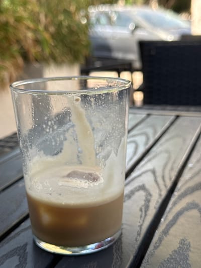 A close-up, eye-level shot shows a partially consumed iced coffee in a clear glass. The drink has a foamy, light brown top and a darker brown liquid base with ice cubes. The glass is sweating, with condensation droplets visible on its surface. The drink is on a dark wooden table with a prominent grain pattern. In the background, out of focus, are green foliage, a light-colored car, and a black chair. The scene appears to be outdoors in a cafe setting, possibly in Campillo Del Moro, Spain, given the context. There are no people visible in the image.