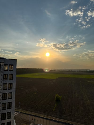 An eye-level, medium shot captures a sunset over a suburban landscape in Kraskovo, Russia. In the foreground on the left, the corner of a modern, multi-story building with a white brick facade and dark-framed windows is visible. The building's windows reflect the sunset's golden light. To the right of the building, a plowed field stretches towards a distant treeline and a hazy city skyline. Another field, a vibrant green, is situated between the plowed field and the treeline. The sun, a bright orb casting a lens flare, hangs low in the sky, surrounded by scattered clouds tinged with orange and yellow. The sky transitions from a pale blue at the top to a muted orange and yellow near the horizon. The environment appears to be an undeveloped or semi-developed area on the outskirts of the city, with agricultural land juxtaposed against modern residential construction. No people are visible in the frame. The overall mood is serene and tranquil, accentuated by the warm hues of the sunset.