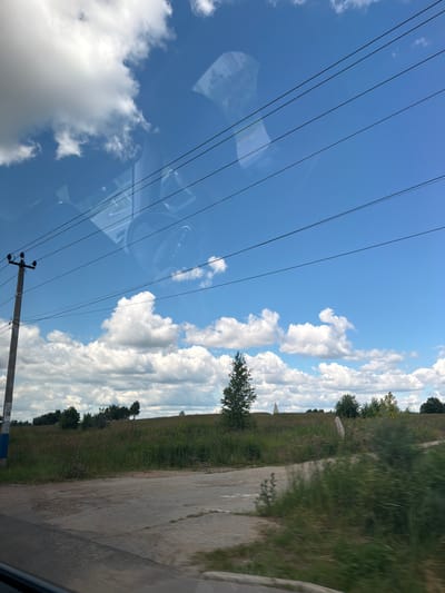The image shows a rural landscape with a clear blue sky dotted with white clouds. In the foreground, a dirt road or path leads into a grassy field. A utility pole with wires stretches across the frame. A lone tree stands in the middle ground, and in the distance, there is a conical structure, possibly a monument or building. The scene appears to be taken from a moving vehicle, as evidenced by the slight blur in the foreground and the reflections on what looks like a car window. The location is specified as Ренесанс Парк Кп, Russia. There are no people or animals visible in the image. The overall impression is one of a sunny day in the countryside.