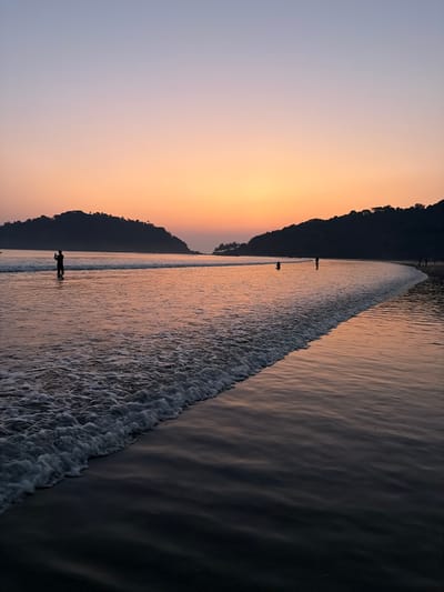 The image depicts a coastal scene during sunset or sunrise at Kolamb, India. The sky transitions from a light lavender hue at the top to a warm orange and pink gradient near the horizon. Two dark, tree-covered landmasses are visible on the distant left and right, with an opening resembling a bay or inlet between them. The ocean occupies the majority of the lower frame, reflecting the sky's colors. A distinct line of white foamy surf runs diagonally across the lower-left foreground. In the water on the mid-left, a solitary individual stands, holding an object (likely a camera or phone) up, facing the horizon. Two other individuals are visible further right, partially submerged in the water. A small group of people are discernible as dark silhouettes along the distant shoreline on the far right. The immediate foreground consists of dark, wet sand exhibiting reflections.