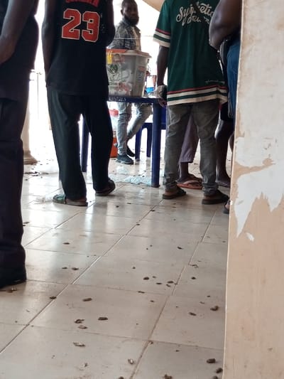 In Ajalli, Nigeria, an indoor or sheltered area is depicted with multiple individuals standing. The floor is tiled with light-colored, possibly off-white, ceramic squares and is littered with numerous dark brown fragments consistent with peanut shells, along with scattered clear plastic bags.
At least five individuals are visible. On the far left, a person's lower left leg is seen, wearing dark trousers and dark footwear. To their right, a male individual stands with his back mostly to the camera, wearing a black sleeveless shirt with "JORDAN 23" printed in red and white, black trousers, and open-toed sandals.
In the mid-ground, slightly left of center, another male individual stands behind a light blue plastic table. He has a beard and wears a light-colored, possibly denim, long-sleeved shirt, light-colored jeans, and dark athletic shoes. On the table is a large, clear plastic bucket with a white lid, containing various items. Underneath the table, at least two blue plastic chairs are visible.
To the right of the table, another male individual stands facing away, wearing a dark green short-sleeved shirt with white and red stripes on the sleeves and bottom hem, and light grey jeans with dark brown open-toed sandals. Further right and partially obscured, the legs of one or more individuals are visible, including someone wearing light purple or pink trousers/skirt and pink flip-flops, and another in blue jeans.
A light-colored column or wall is partially visible on the far left, and a beige-colored wall with peeling paint or plaster is on the far right. The individuals appear to be standing, possibly waiting or engaged in an informal gathering.