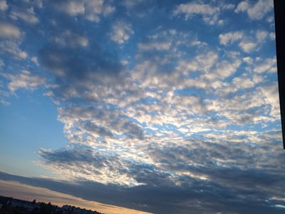 This is a photograph of a cloudy sky taken during sunset. The clouds are varied, with some appearing dark and heavy, while others are illuminated by the setting sun, creating a striking contrast of light and shadow. The sky transitions from a deep blue to warmer, golden hues near the horizon, suggesting the end of the day. In the upper portion of the image, the silhouettes of buildings and trees are visible against the sky, indicating a populated area. The image is rotated, giving the impression of looking up at the sky from an unusual angle, or perhaps reflecting the sky in a body of water. The overall mood is peaceful and serene, with the dramatic interplay of light and cloud formations adding a touch of awe. The location is specified as Shostka, Ukraine.