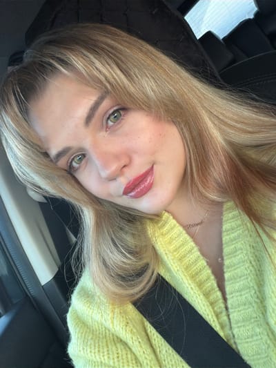 A young adult female is depicted in a close-up portrait, positioned inside a vehicle. Her blonde hair with bangs frames her face, complementing her green eyes. She has light skin with visible freckles on her cheeks and is wearing glossy pink lipstick. A slight smile is present as she looks directly towards the camera. She is wearing a bright yellow knitted cardigan or sweater. A thin gold necklace with a small pendant (partially legible as a word, possibly "Valeria") is visible around her neck. A black seatbelt diagonally crosses her chest. The background consists of dark, likely leather, car upholstery and a glimpse of a window in the upper right portion of the frame. There is no visual information within the image to determine the specific location within the city of Азьмушкино, Russia.