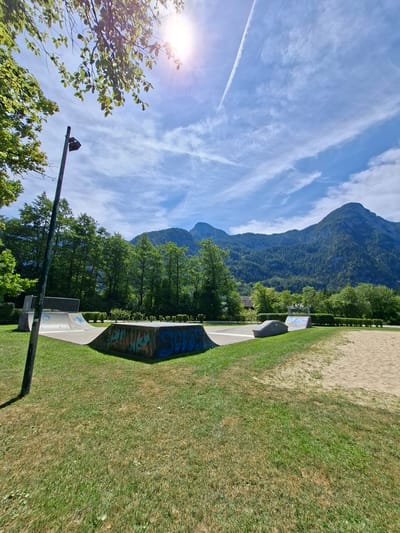 The image depicts a skatepark in Obertraun, Austria, bathed in bright sunlight under a partly cloudy blue sky. In the foreground, a green grassy area transitions into a sandpit on the right. The skatepark features several concrete ramps, one of which is covered in colorful graffiti. A tall, dark lamppost stands to the left of the park. Behind the skatepark, a dense line of lush green trees forms a backdrop, beyond which majestic, tree-covered mountains rise, their peaks partially obscured by the sky. A contrail from an aircraft is visible streaking across the sky. The sun, positioned in the upper left quadrant, casts a lens flare onto the image.
