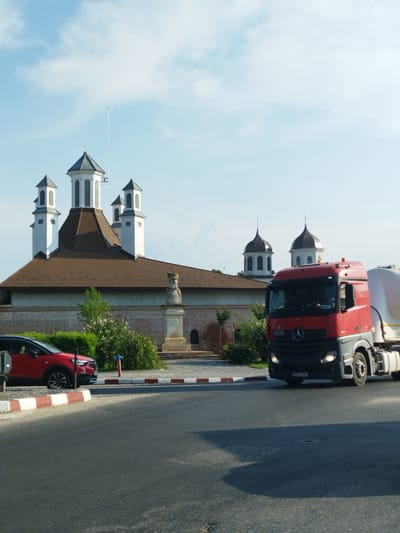 The image depicts the exterior of the Cantacuzino Manor in Potlogi, Romania, bathed in daylight under a partly cloudy sky. In the foreground, a grey asphalt road curves to the right, bordered by a red and white striped curb. A red compact SUV is parked on the left side of the road. To the right of the SUV, a large red tanker truck is driving on the road, its front visible.
The Cantacuzino Manor is characterized by its distinctive architecture. The main building features a large, steeply pitched brown roof and a red brick lower wall. Several white towers with conical roofs and dark spires rise from the building, some topped with smaller spires. A statue of a robed figure wearing a crown stands on a pedestal in front of the manor, partially obscured by greenery. Lush green bushes and trees surround the base of the manor and the statue. The overall scene suggests a historic and well-maintained site on a clear day.