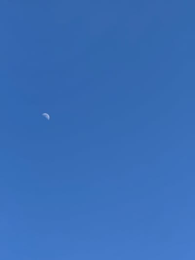 The scene depicts a clear, uniform blue sky. A crescent moon, exhibiting a distinct illuminated curve and discernible surface features, is visible in the upper-left portion of the frame. The sky is entirely clear, with no clouds, people, or other objects present. The location is the Cherwell District, United Kingdom.