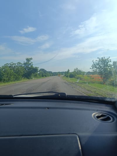 The image shows a view from the interior of a vehicle, looking out at a paved road stretching into the distance under a bright, partly cloudy sky. The road is flanked by lush green trees on the left and a field of red poppies and green vegetation on the right. A yellow bus is visible in the distance on the road. A faint contrail is visible in the sky. The foreground shows the dashboard of the vehicle, with part of the windshield wiper visible. The scene appears to be a rural setting, likely on the outskirts of Okny, Ukraine, given the presence of poppies, which are common in the region, and the general landscape. There are no people visible in the image.