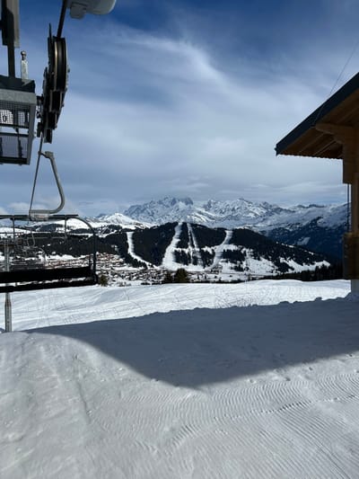 The image presents a panoramic winter landscape from a ski resort in Villard-sur-Doron, France, under a partially cloudy blue sky. In the foreground, snow-covered ground exhibits numerous ski tracks and a distinct dark shadow cast across its surface. The snow also shows textured ripple patterns. The upper-left quadrant features the intricate metal structure of a ski lift mechanism, including cables, pulleys, and a suspended empty ski chair labeled "803 Doppelmayr." To the far right, the corner of a building with a dark, sloped roof and exposed wooden eaves is visible. The midground encompasses a valley with a village comprised of multiple buildings, identifiable by their reddish-brown roofs, situated amidst dark evergreen trees. Several wide, groomed ski slopes with visible tracks descend through the forested mountain. The background is dominated by an extensive range of rugged, snow-capped mountains. No individuals are present in the scene.