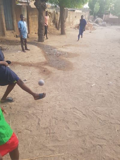The image depicts an outdoor scene in Dutsinma A, Nigeria. The ground is sandy and uneven. Several people are present, with two barefoot men standing near a dilapidated structure made of mud bricks. Another man in blue clothing is lying on the ground further away. In the foreground, a person wearing shorts and sandals is kicking a white ball. The overall impression is a candid shot of everyday life.