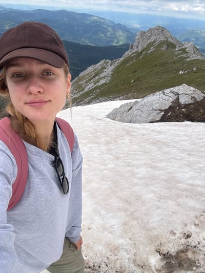 A young woman wearing a brown baseball cap, a grey sweatshirt, and khaki pants stands on a snow-covered mountain slope, taking a selfie. A pink backpack is visible on her shoulders. She has a blonde braid and is wearing a gold hoop earring. Sunglasses are clipped to her sweatshirt. The background shows a vast mountainous landscape with green slopes and rocky peaks, under a cloudy sky. The presence of snow suggests a high-altitude location, potentially in the mountains near Jošanica, Montenegro, though this cannot be definitively confirmed from the image alone.