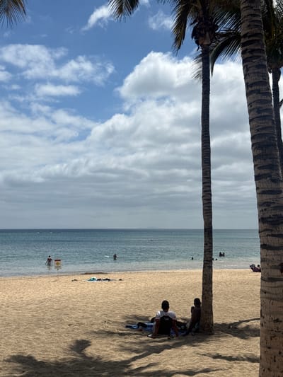 The image shows a beach scene in Arrecife, Spain. Two palm trees frame the view. The beach is sandy, with a visible tide line. People are scattered along the shoreline and in the water. A warning sign is posted near the water's edge. In the foreground, two individuals are seated beneath the shade of a palm tree. Clothing and personal items are spread out around them. The sky above is partly cloudy.
