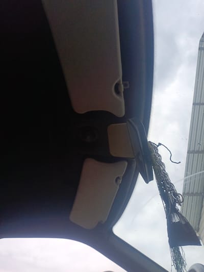 The image shows the interior of a vehicle, looking upwards towards the windshield and the car's roof lining. On the left, a beige sun visor is visible, partially obscuring the dark roof. In the center, a black rearview mirror is attached to the windshield and hangs down. A decorative item made of chains and a black fabric fringe is hanging from the mirror. Through the windshield, a cloudy sky and a building with vertical siding are visible. The photograph was taken in the city of Gil'deevo, Russia.