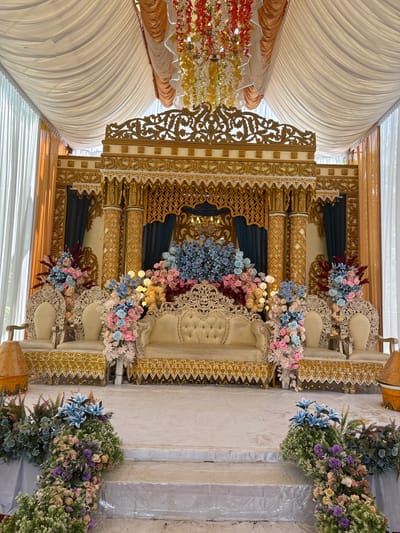 This image depicts a decorated wedding stage at an outdoor venue in Kruengseupeng, Indonesia. The stage features an ornate golden backdrop with intricate carvings and decorative fabric draping. A cream-colored tufted sofa and two armchairs are arranged centrally on the stage, adorned with numerous artificial flower arrangements in shades of blue, pink, and white. The stage is elevated by a white carpeted platform with steps leading up to it, also decorated with floral arrangements. The surrounding area appears to be a covered outdoor space, with sheer white curtains and some orange drapery visible. The overall impression is one of a lavish and festive setting for a wedding ceremony. There are no people visible in the image.