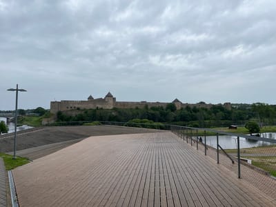 The image depicts a wide wooden promenade in the foreground, extending towards a body of water. On the opposite bank of the water, a large, historic stone fortress with multiple towers is prominently visible. The fortress is situated on a hill and is surrounded by lush green trees. The sky above is overcast with heavy clouds. In the background, a bridge can be seen crossing the water. The overall scene suggests a view of the Narva Castle in Narva, Estonia, from a waterfront park or viewing area.