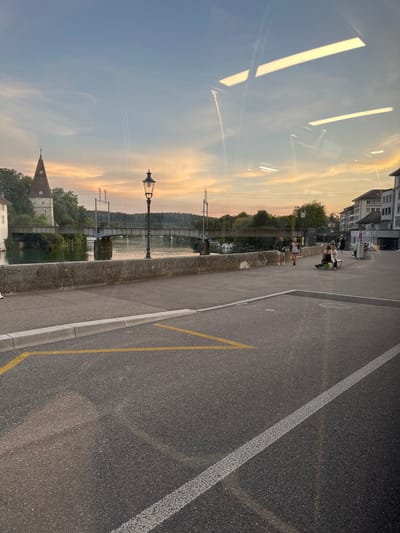 The image depicts an urban riverfront scene in Solothurn, Switzerland, at dusk. The view is across the Aare River, with the Wasserturm (Water Tower) and a railway bridge visible on the left.
In the foreground, a section of dark asphalt pavement occupies the lower third, featuring a solid white lane line on the right and yellow angular markings on the left. A grey concrete curb delineates the road from an elevated, paved pedestrian walkway.
Along the walkway, a stone retaining wall runs parallel to the river. A dark, ornate streetlamp with a single light fixture stands near the center of the frame. The Aare River flows behind the wall, its surface reflecting the ambient light. A multi-span railway bridge, supported by concrete piers, crosses the river, carrying tracks and overhead electrical lines. On the far left bank, the historic Wasserturm, a stone tower with a distinctive brown conical roof, is partially obscured by trees. Lush green foliage lines both riverbanks. Several small boats are moored along the river, predominantly on the right side of the bridge.
On the pedestrian path, two individuals in dark athletic clothing are jogging or power-walking towards the right. Further along the path, near the right edge of the frame, a small group of people is gathered; one person is seated in a mobility device, and another is seated on a public bench. Buildings with multiple stories and standard urban architectural features line the far right bank.
The sky transitions from light blue directly overhead to hues of orange, pink, and yellow near the horizon, indicative of sunset. Faint white contrails are visible in the upper sky. Two distinct, elongated horizontal reflections of interior lighting, likely fluorescent fixtures, are visible on the surface of the image, suggesting the photograph was taken through glass.