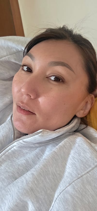 A close-up, angled view of a woman with brown eyes and dark hair pulled back. She wears a light gray zip-up hoodie. Her face is well-lit, revealing clear skin with some freckles. The background suggests an indoor setting with a wooden door visible in the lower left. The location in the city of Lensk, Russia, is not discernible from the image.