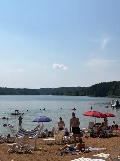 This is a medium-shot, vertically oriented photograph taken on a sunny day at a beach resort located in the Ozyaritska-Slabadski rural council, Belarus. The scene depicts a beach with people relaxing and swimming. In the foreground, several beach chairs are set up on the sand, some occupied by people. Beach umbrellas, one red and one purple, provide shade. The water appears calm, and several individuals are visible swimming or wading. Further out in the water, there are inflatable structures. The background features a densely forested hillside, characteristic of the Belarusian landscape. The sky is a clear blue with a few wispy clouds, suggesting fair weather. The overall mood is one of leisure and recreation on a warm day. No visible text is present in the image.