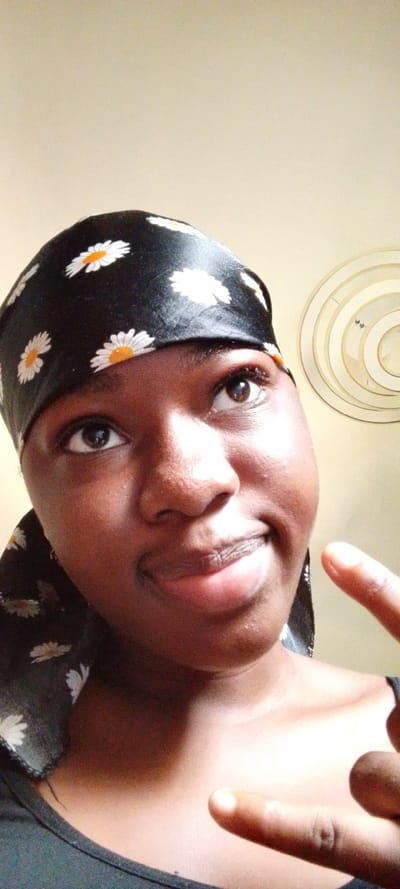 A close-up portrait shot captures a dark-skinned individual, likely female, framed from the chest upwards. The individual wears a black headwrap patterned with multiple white daisies, each featuring an orange center. Their eyes are directed upwards and slightly to the right of the frame. A subtle smile is present, with lips slightly parted. The individual's right hand is visible in the lower right quadrant, with the index finger extended and pointing towards the lower left of the frame. The visible clothing consists of a dark-colored shirt. The background features a plain, light-colored wall. In the upper right quadrant, a circular, multi-layered light fixture or decorative element is visible. The scene is situated in the city of Itu, Nigeria.