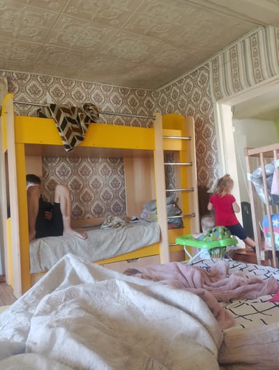 In a room with patterned wallpaper and a textured ceiling, a bunk bed with a bright yellow frame dominates the scene. On the lower bunk, a young person in black shorts and a dark shirt is seated, looking down at a phone. A patterned blanket is draped over the upper bunk. To the right of the bunk bed, a young girl in a red shirt and dark pants stands in front of a wooden crib. A baby is seated in a green and blue baby walker nearby. The room appears to be a bedroom, likely in a home. The lighting suggests it is daytime. No text is visible in the image. The overall impression is a domestic scene with children and a young adult engaged in relaxed activities.