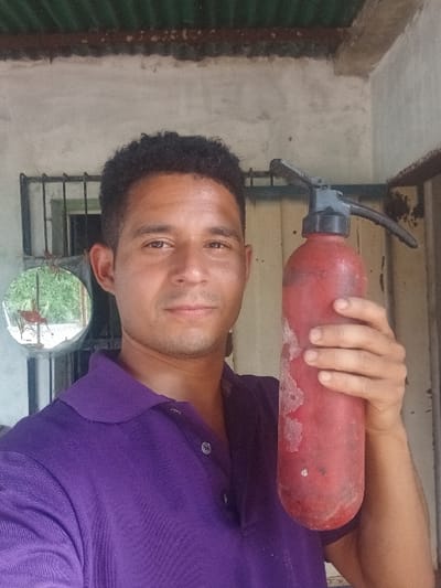 An adult male is depicted in a close-up, chest-up selfie-style photograph taken in Cujisal, Venezuela. He has short, dark, curly hair and medium-toned skin, looking directly at the camera with a neutral expression. He wears a purple polo shirt with three visible buttons. The male is holding a red, cylindrical fire extinguisher upright in his right hand, positioned in front of his torso. The fire extinguisher appears worn, with white residue or discoloration visible on its surface, and features a black handle and nozzle mechanism.

The background consists of a light grey, unpainted concrete or stucco wall. To the left, a dark-framed window with security bars is visible, behind which a circular mirror reflects outdoor greenery and red furniture. At the top of the frame, a section of a dark green corrugated metal roof is present. To the right of the male, a vertical section of a lighter, possibly wooden or painted, discolored wall or door frame is visible. Natural lighting illuminates the scene.